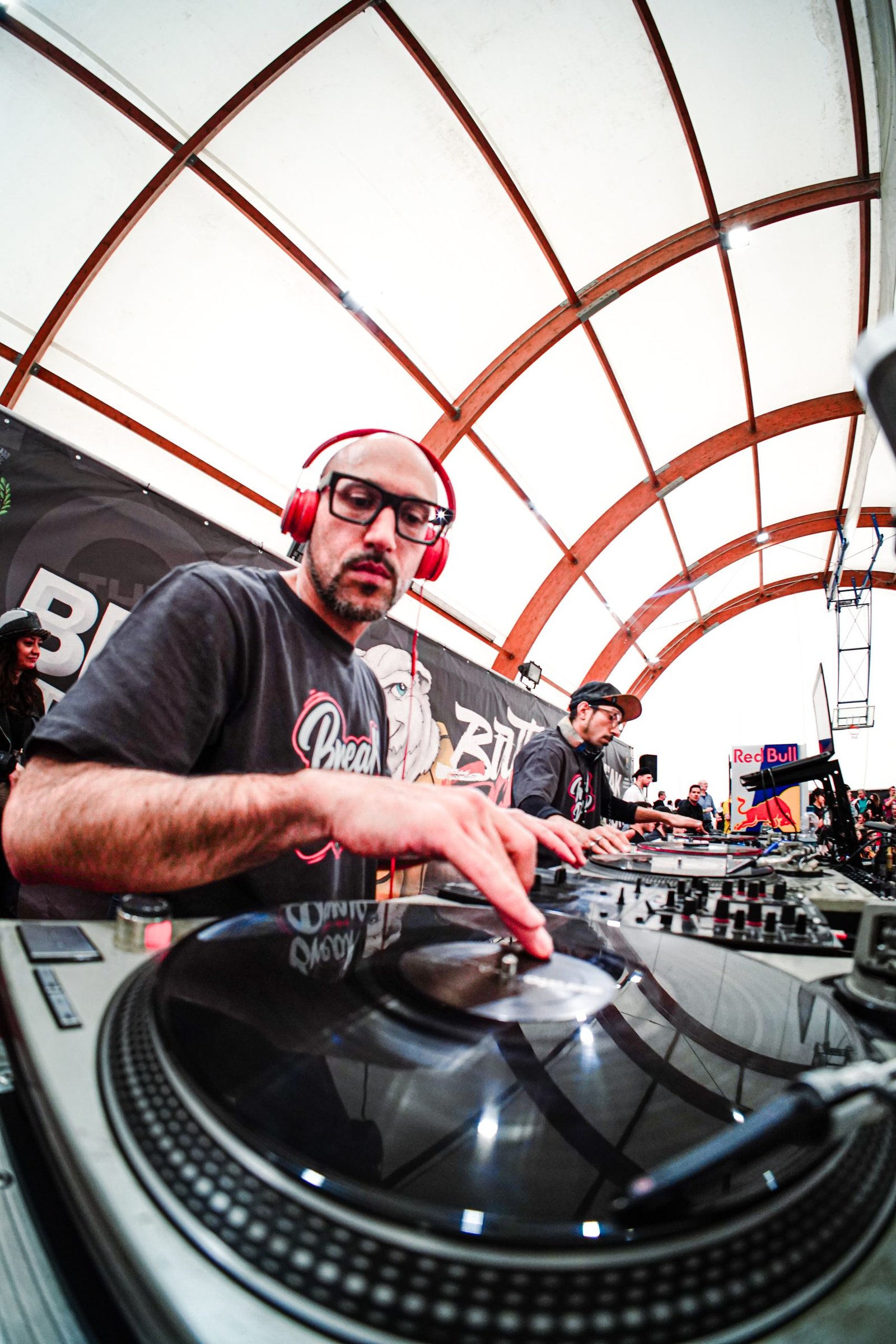 DJ con cuffie rosse e occhiali in consolle durante evento Sparta Bandits Crew, struttura ad arco e banner Breakdance